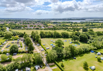 Aerial view of South Lytchett Manor Holiday Park surrounded by green fields and trees, with caravans, motorhomes, and tents pitched in a peaceful Dorset countryside setting near Poole.