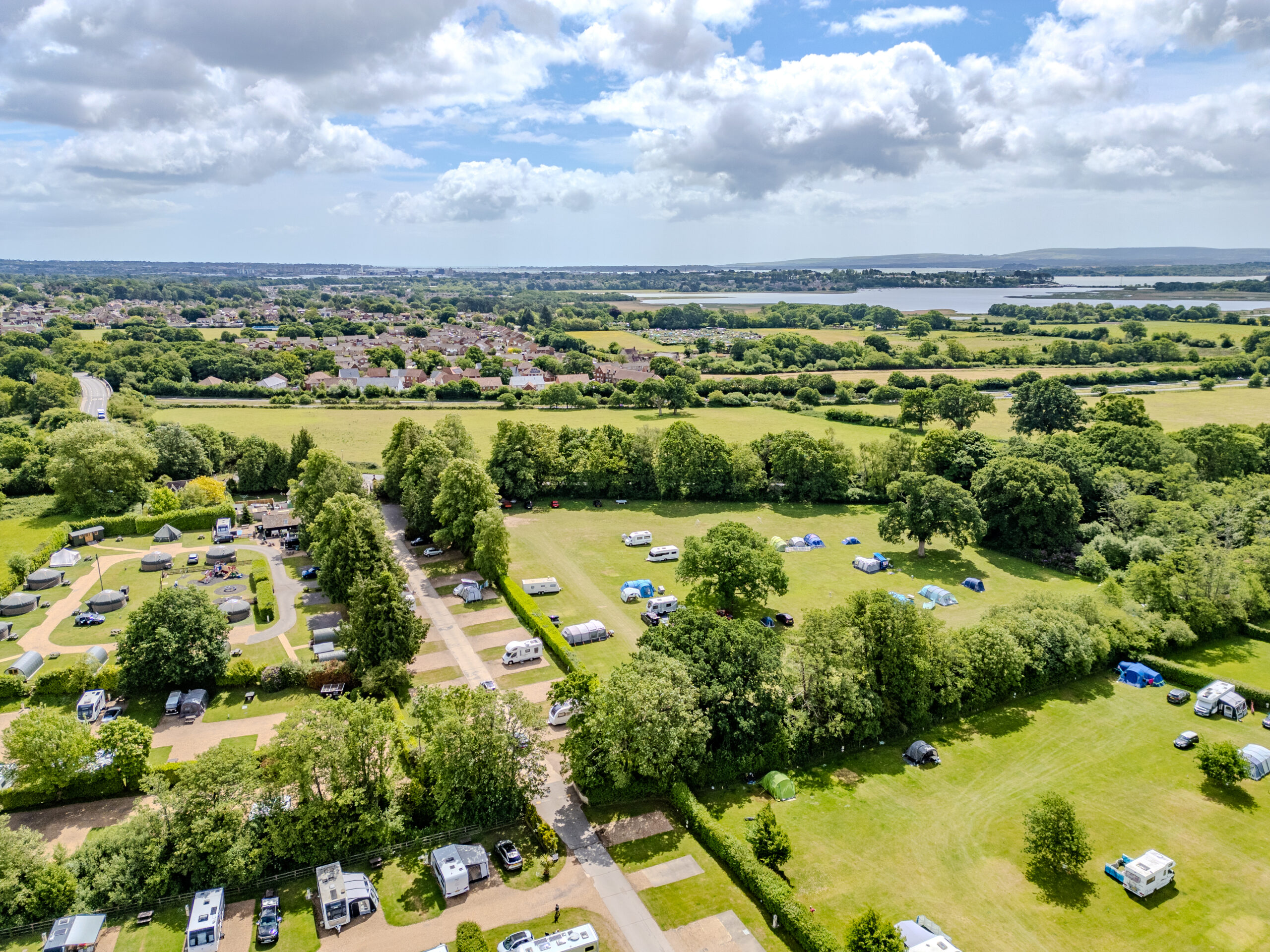 Aerial view of South Lytchett Manor Holiday Park surrounded by green fields and trees, with caravans, motorhomes, and tents pitched in a peaceful Dorset countryside setting near Poole.